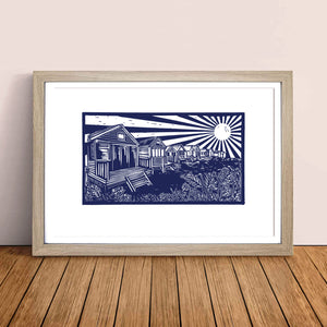 MarcoLooks framed print of the Beach Huts at Mudeford, printed in blue ink. Taken originally from a lino cut print. The print is pictured in a wooden frame, resting against a pale peach wall, on a oak floor board.